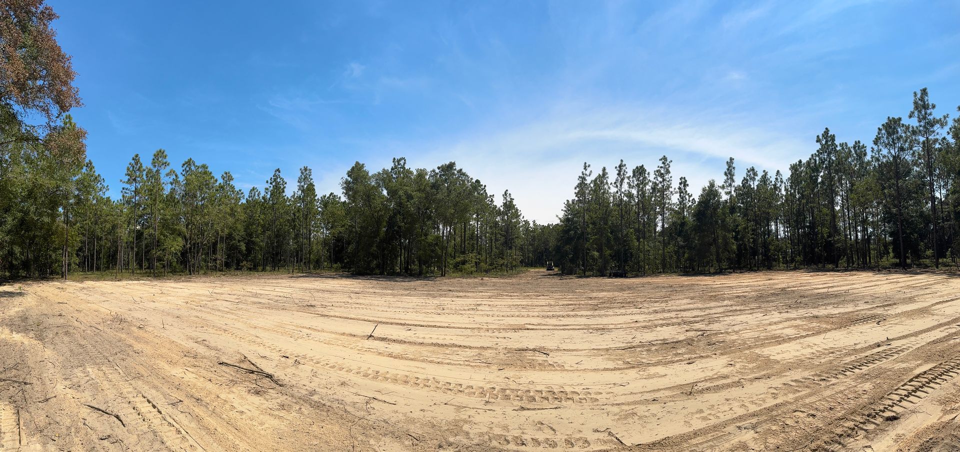 A cleared sandy lot with tire tracks, with a tree line against a blue sky.