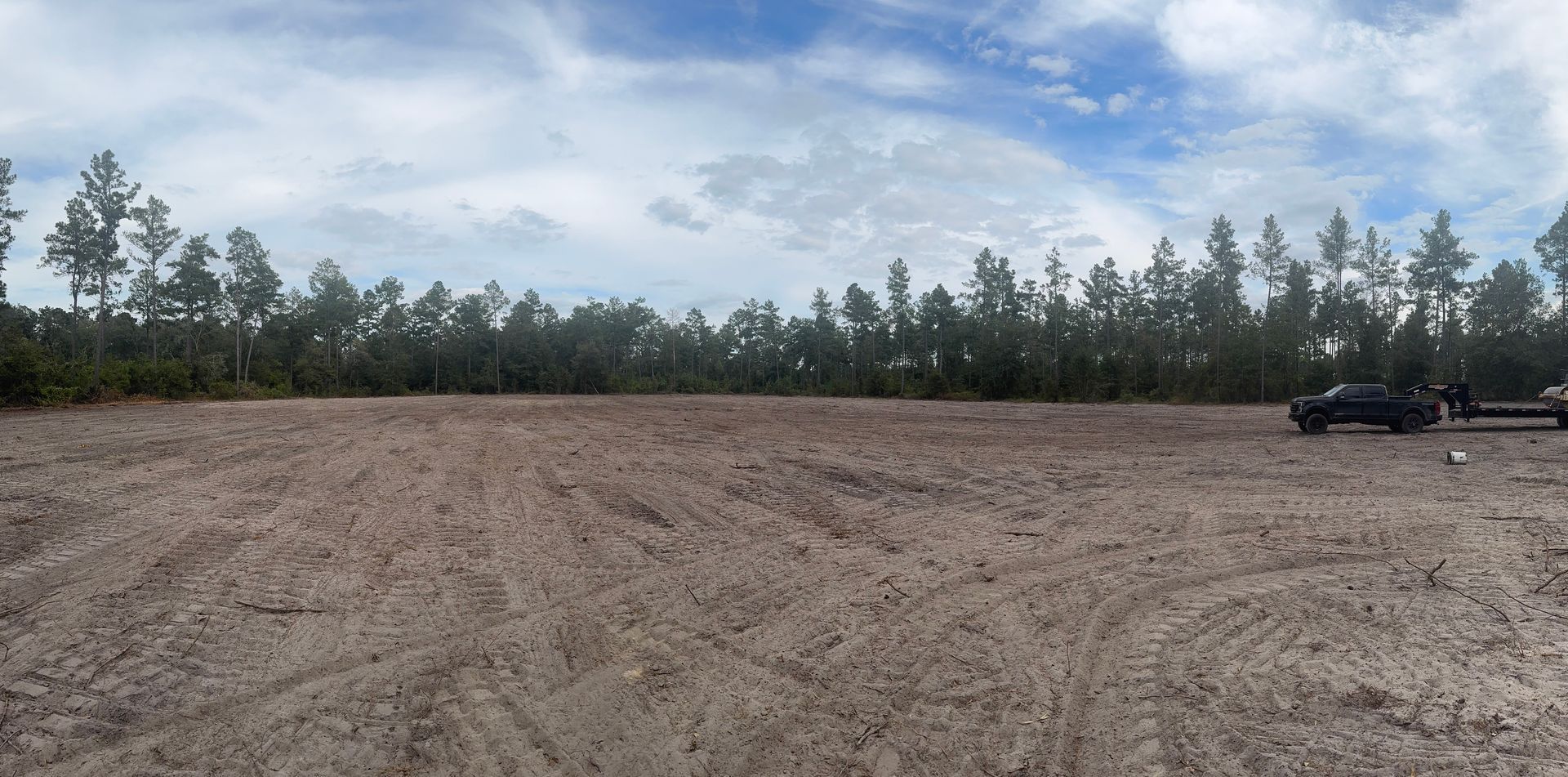 A cleared dirt field with a treeline in the background and cloudy sky. A truck with a trailer is on the right.