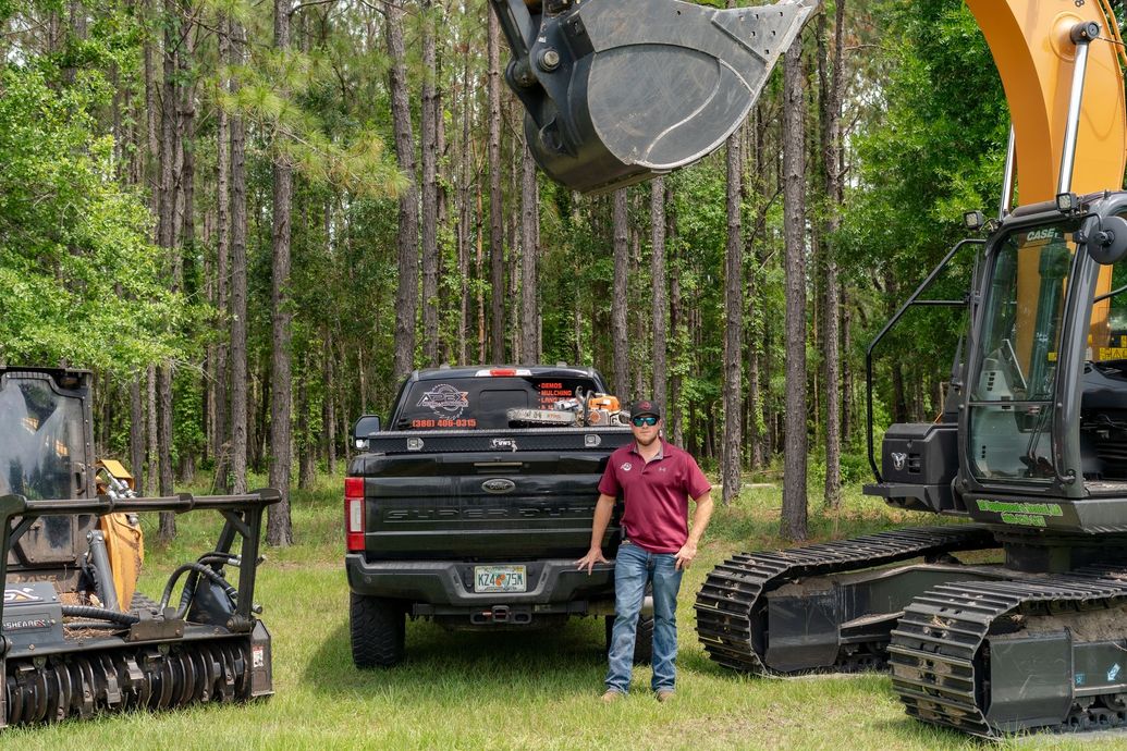 Man standing by truck and heavy equipment in a wooded area.