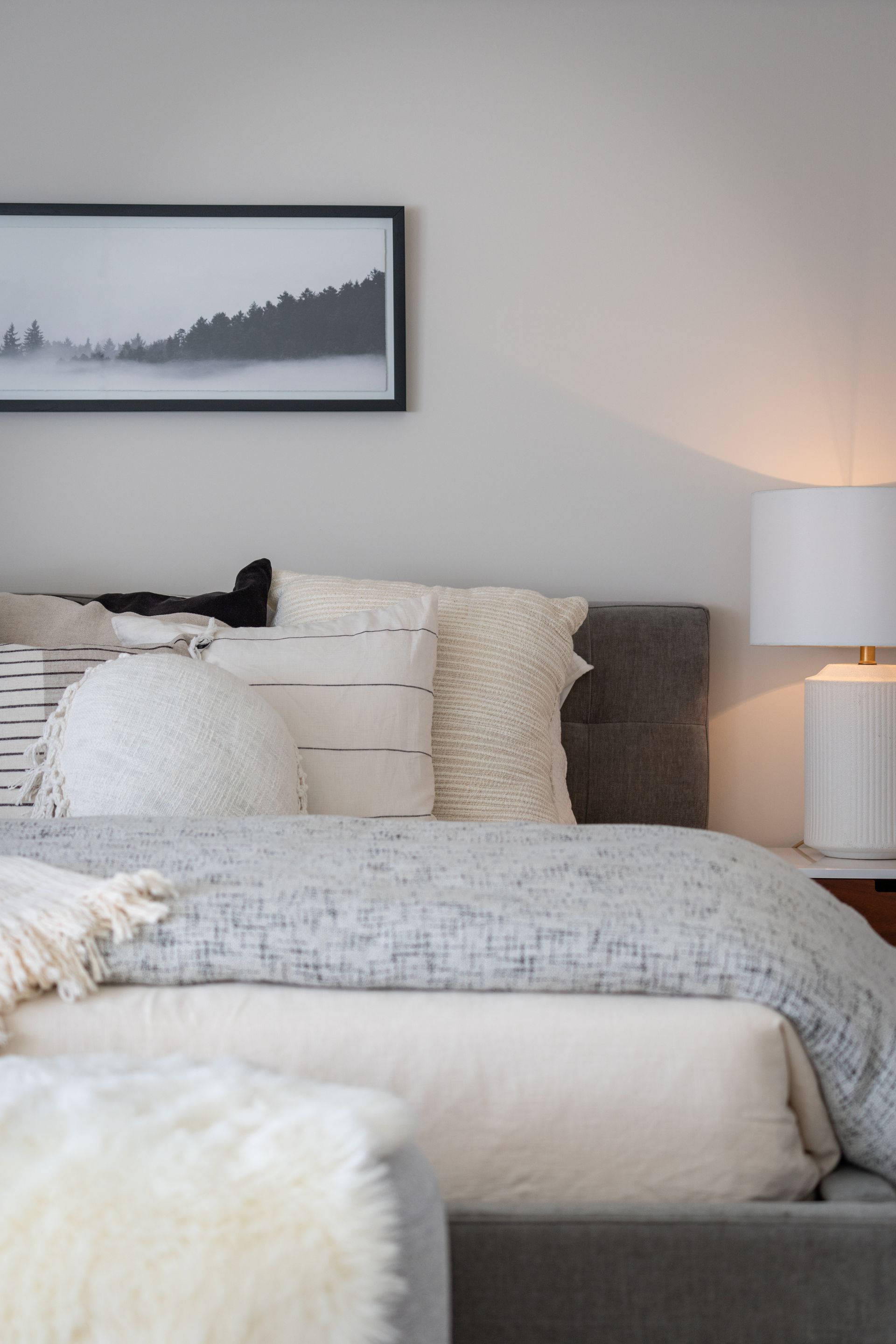 Cozy bedroom with a neatly made bed, neutral bedding, framed landscape print, and bedside lamp