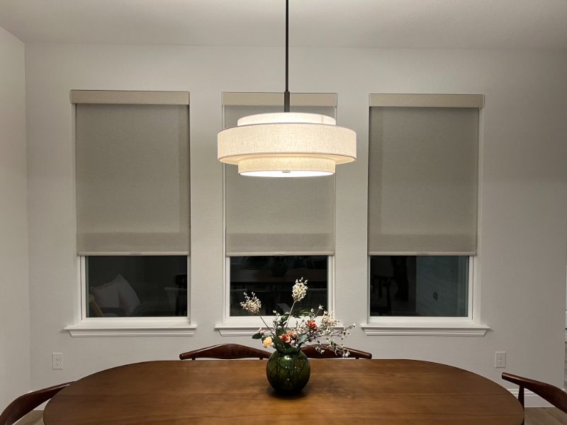 A dining room table sits in front of three windows with motorized window shades. 