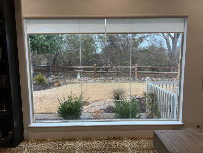 A large window overlooking a wooded backyard has a white roller shade. 