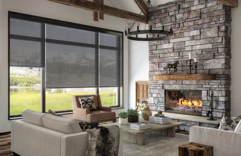 A mountain cabin living room has a large stone fireplace and black roller shades on the windows. 