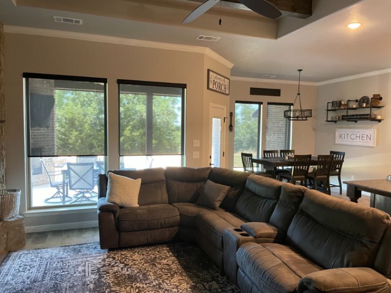 A living room with a large brown couch has black solar shades on the windows. 