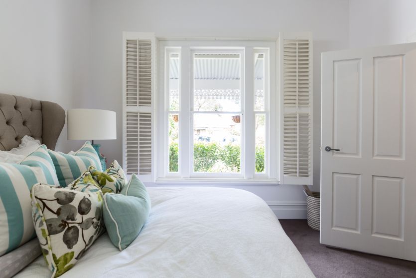 A bright sunny white bedroom has a bed and white shutters on an open window. 