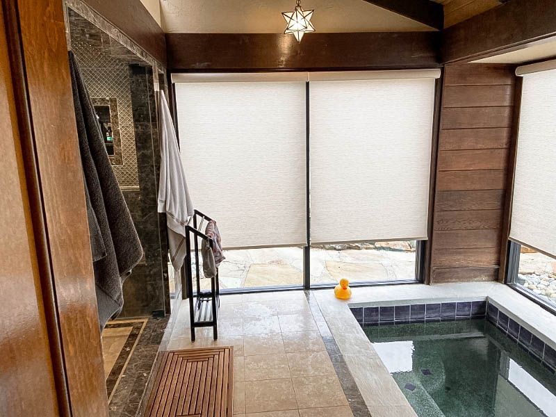 A room with a hot tub has roller shades on large windows. 