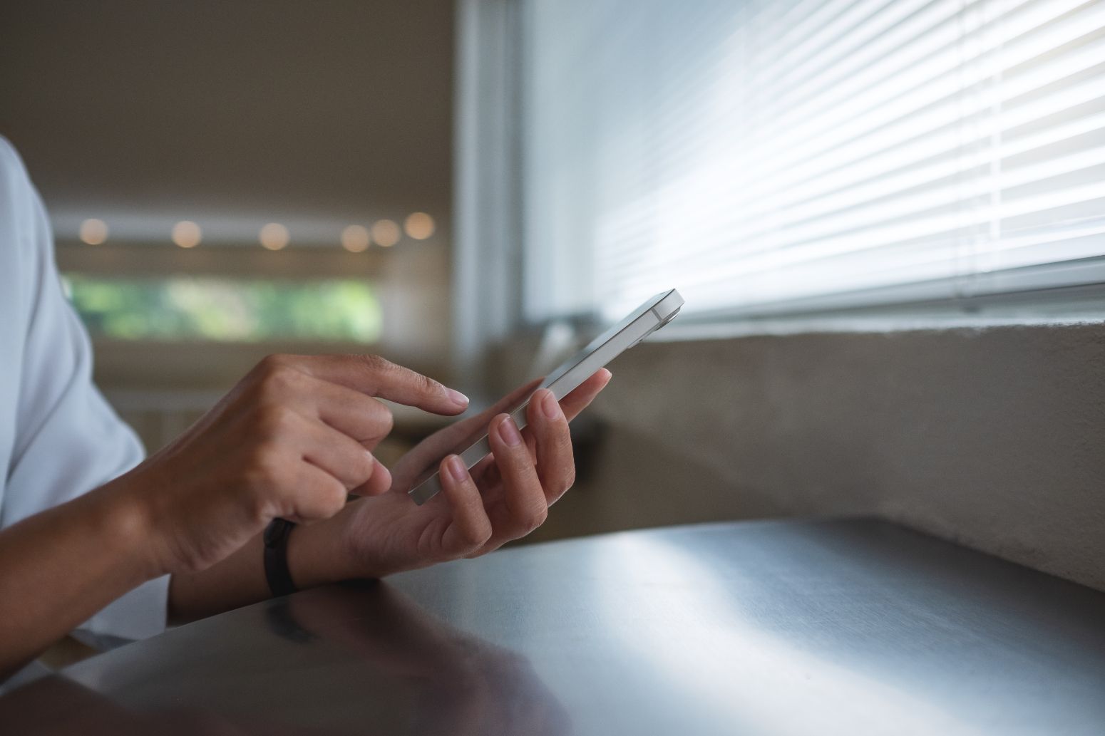 Person using a smartphone by a window; hands tap the screen, natural light.