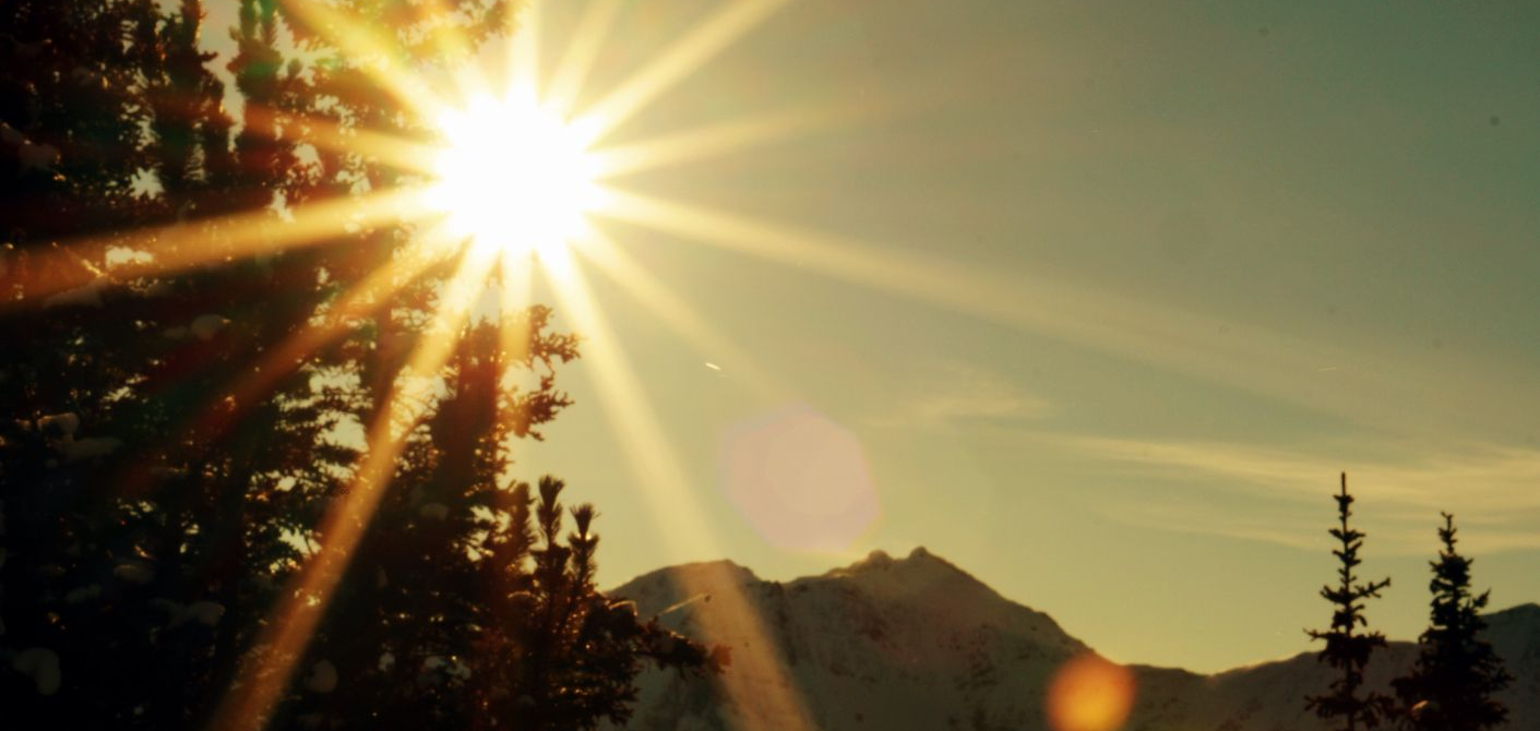 Bright sunburst over mountain range, trees in the foreground. Golden light.