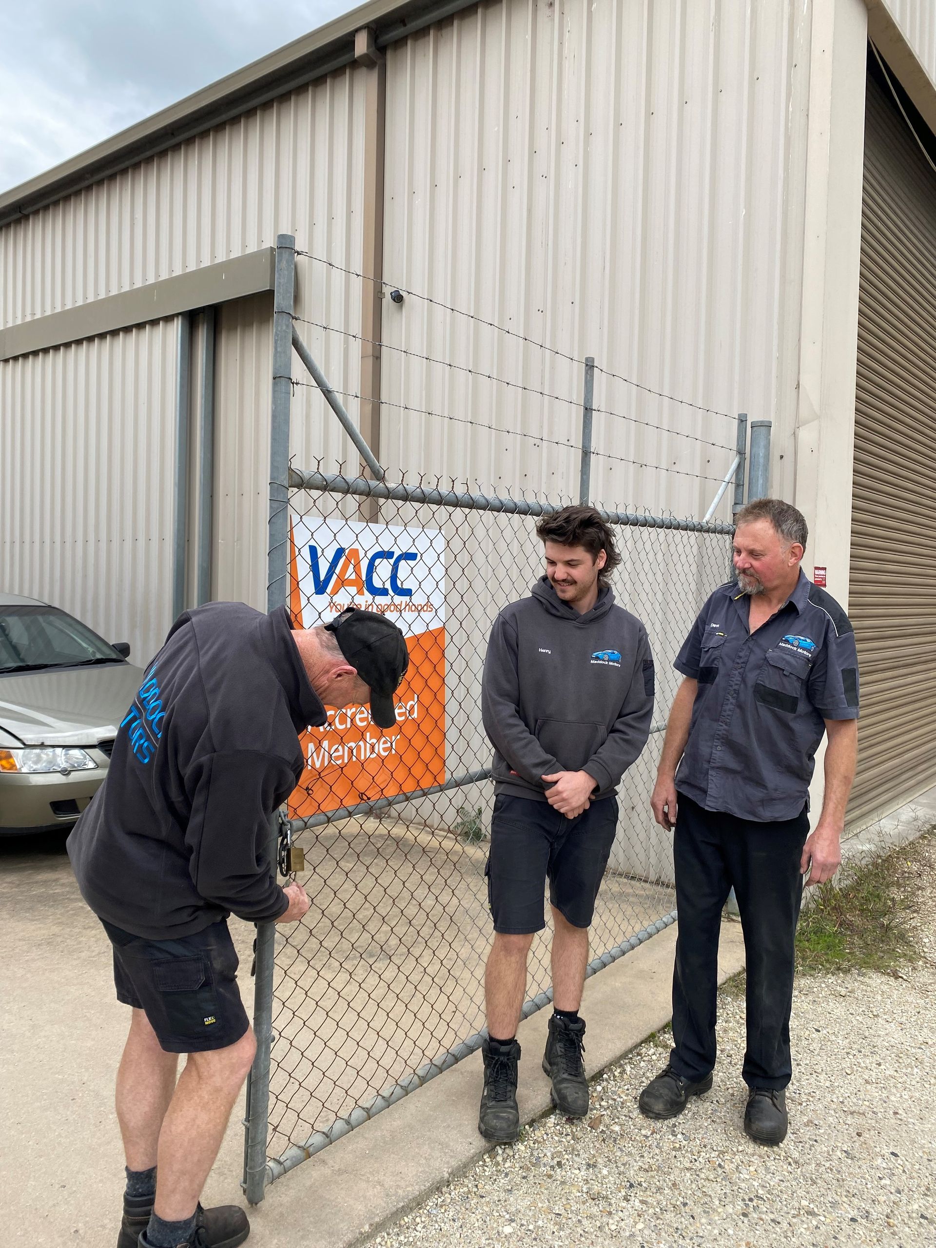 Three men are standing next to a chain link fence in front of a building — Auto Air Conditioning in Wodonga, VIC
