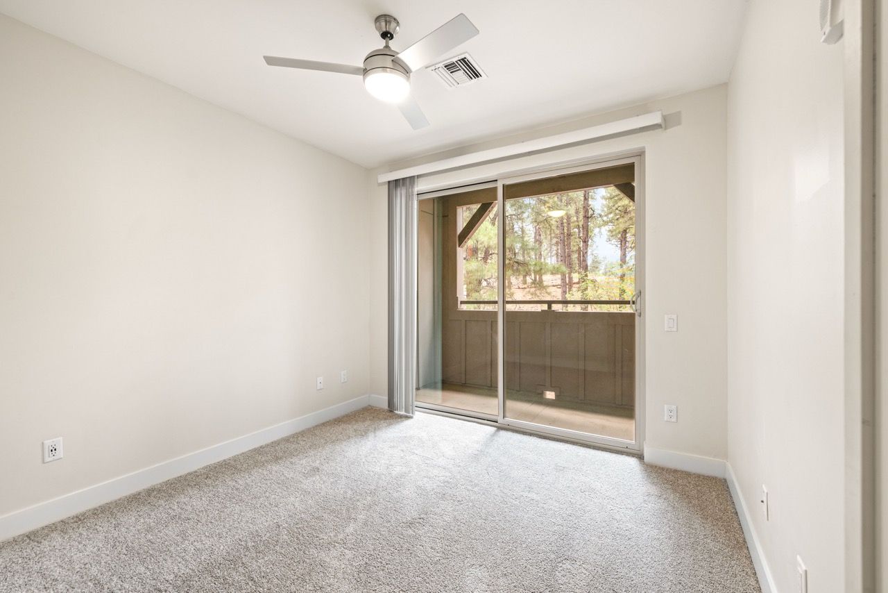 Empty bedroom with beige walls, carpet, sliding glass door to balcony, and a ceiling fan.