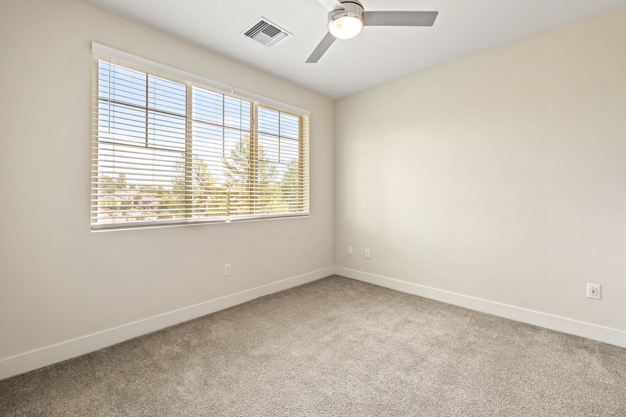 Empty bedroom with beige walls, carpet, a large window with blinds, and a ceiling fan.