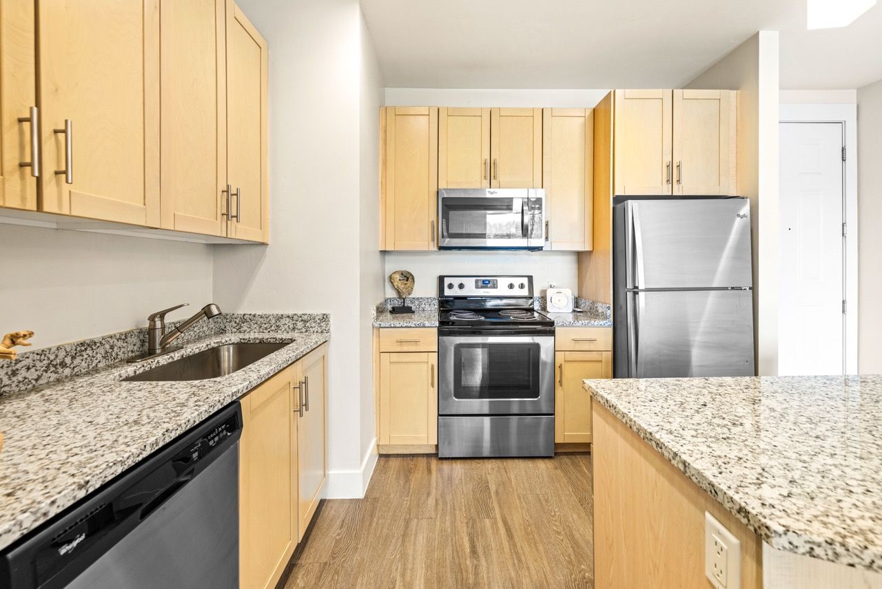 Modern kitchen in apartment with granite countertops, light wood cabinets, and stainless steel appliances.