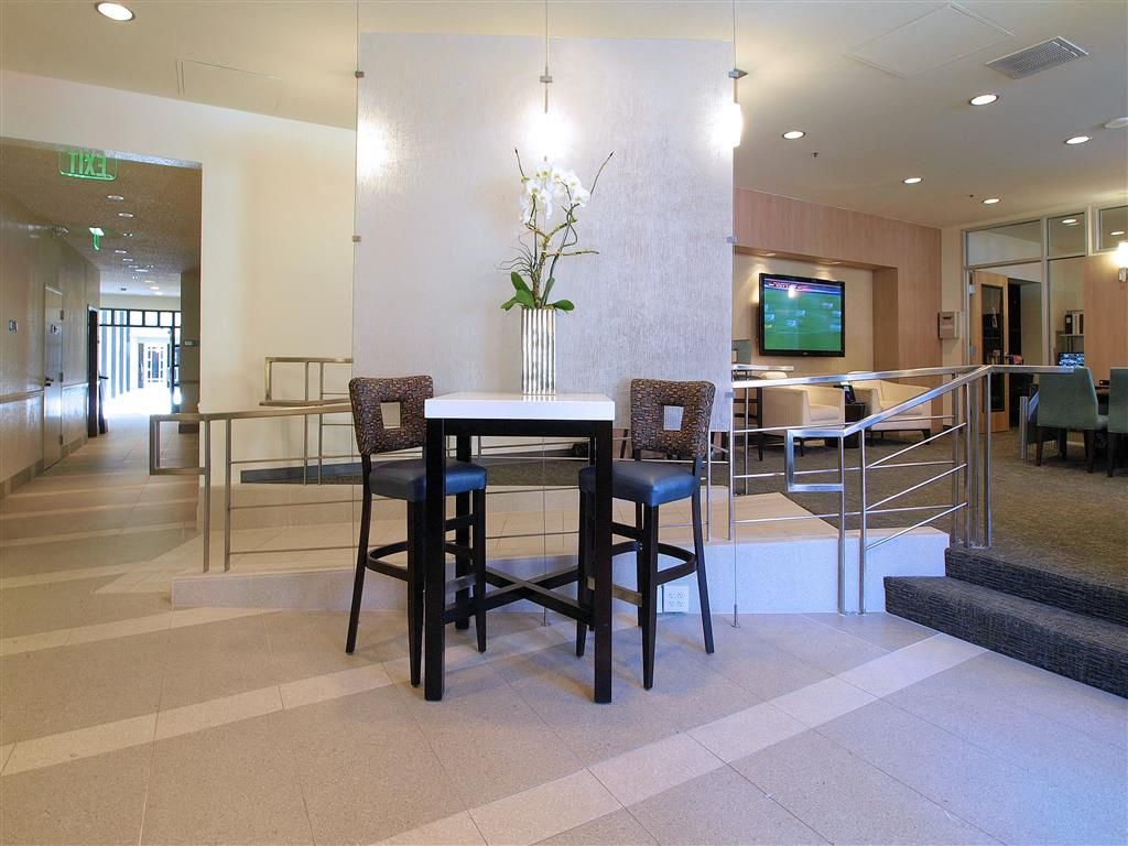 Interior of a modern apartment lobby with a tall table, two stools, a flower centerpiece, and a wall-mounted TV.