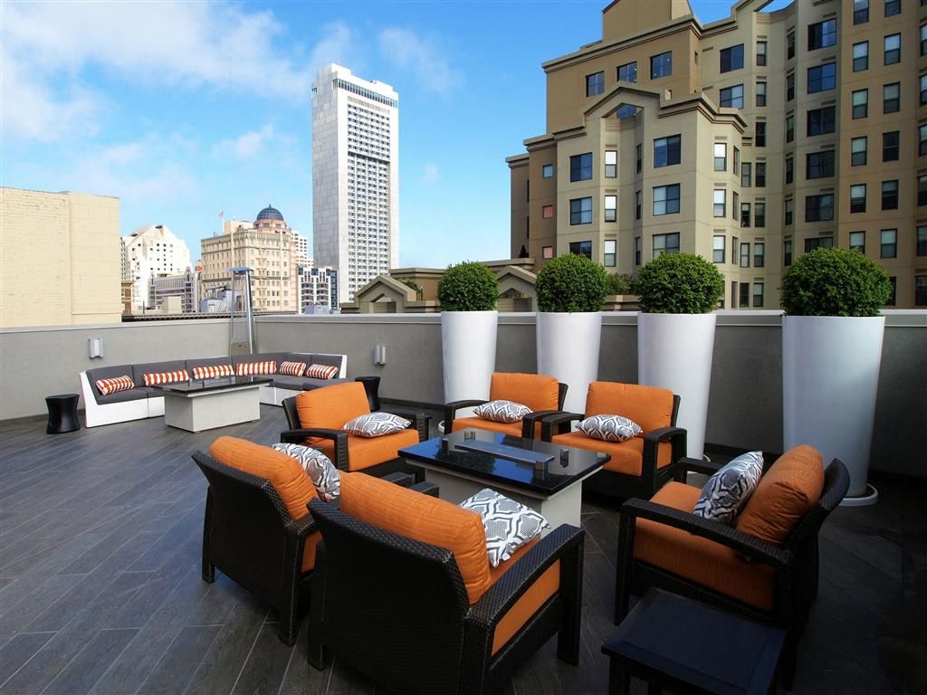 Rooftop community terrace with orange-cushioned lounge seating and planters, city skyline in background.