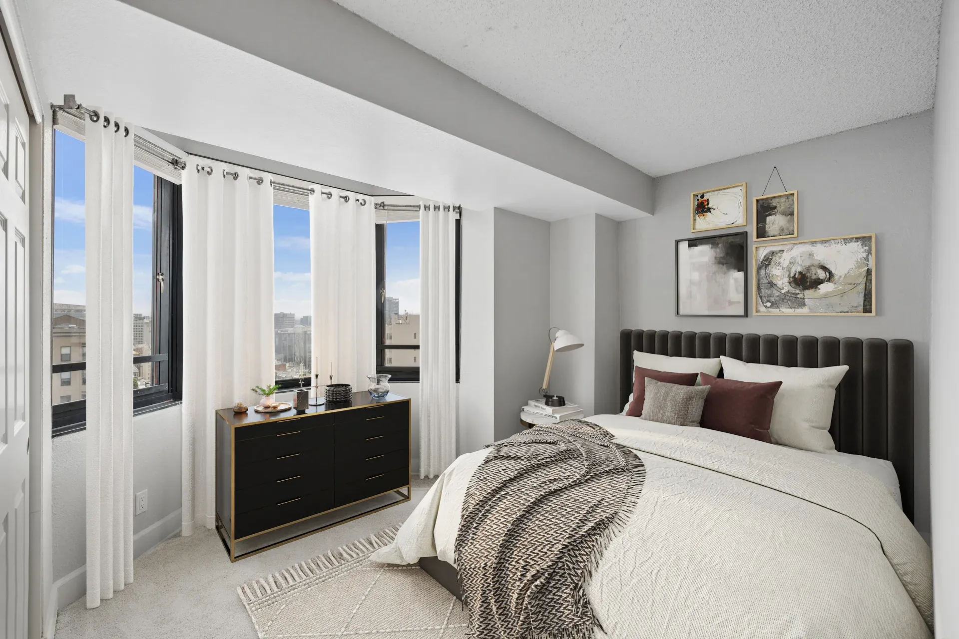 Modern bedroom in an apartment with large windows, sheer white curtains, a dark upholstered headboard, and a dresser.