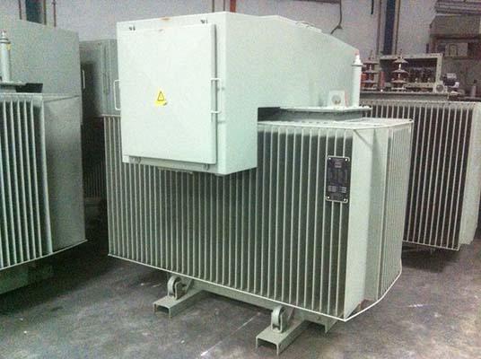 Grey industrial electrical transformer with warning symbol on a light-colored enclosure.