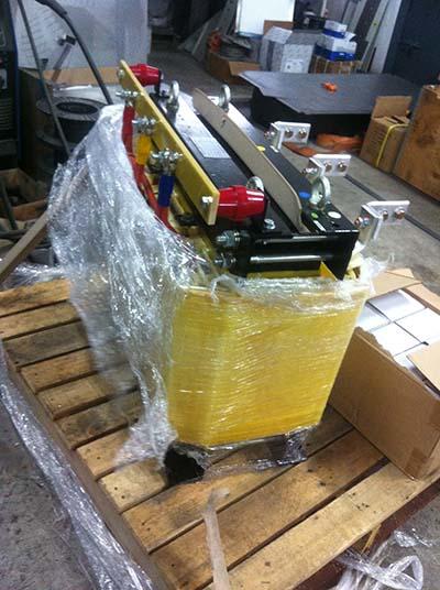 Machine wrapped in plastic on a wooden pallet. The machine is yellow, black, and red. Indoors.
