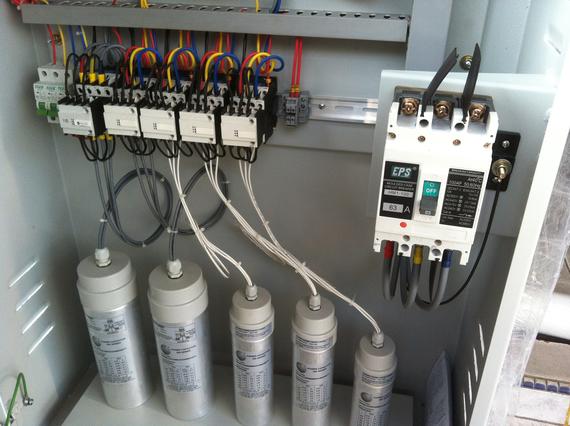 Electrical panel with capacitors, circuit breakers, and wiring in a metal enclosure.