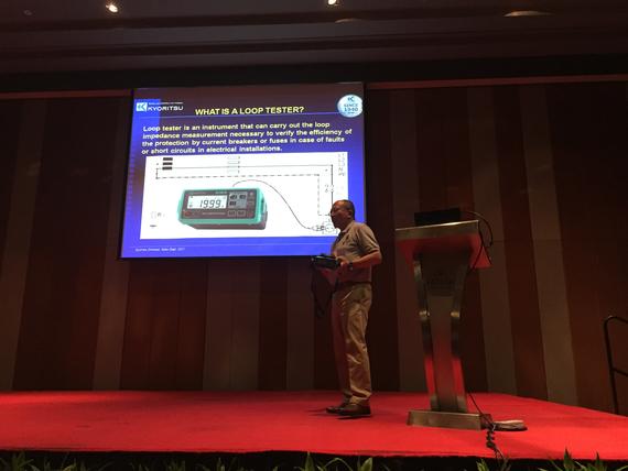 Man presenting on stage; slide shows a loop tester with a diagram. Red carpet, orange backdrop.