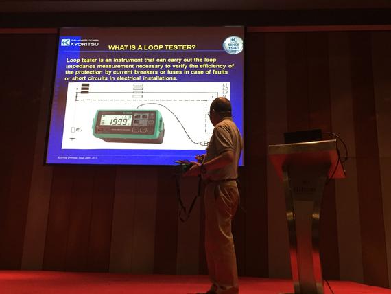 Man presenting loop tester information on screen; dark room, green equipment on display.