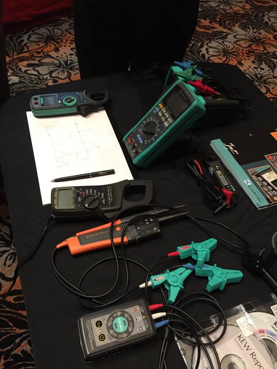 Various multimeters and electrical testing tools on a black surface with a notepad and pen.