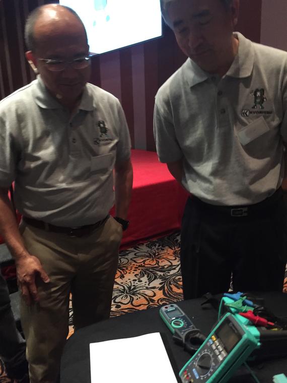 Two men looking at equipment on a table. One wears glasses and a gray polo shirt; the other also wears a gray polo.