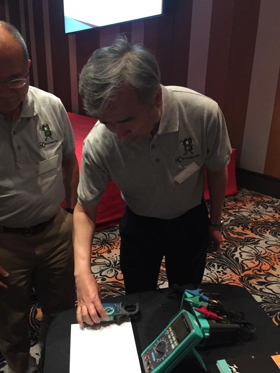 Two men examining electrical equipment on a table; one leans close, touching a device, indoors.