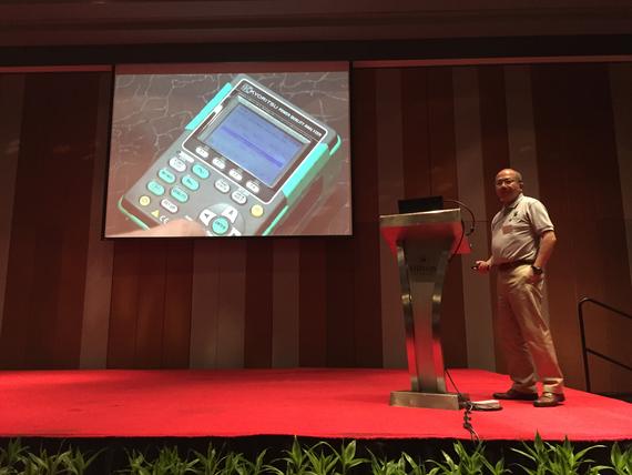 Man presenting, on a red stage, with a large screen displaying a close-up of a green device.