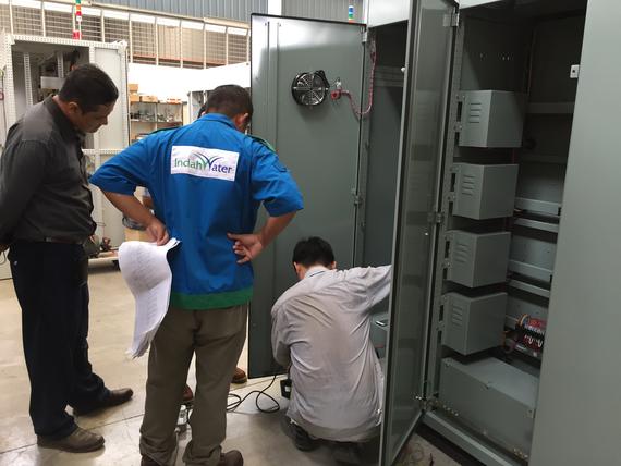 Three men inspecting electrical panel; one is kneeling, another looking on with paperwork. Gray metal cabinet.