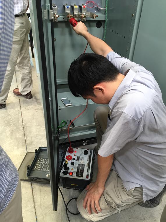 Man testing electrical panel with a handheld device. Indoors, gray panel, wires, and a second man nearby.