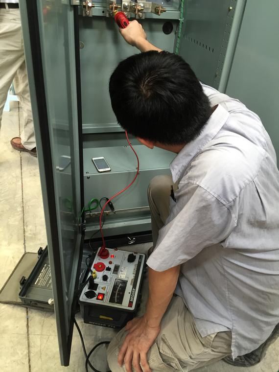 Man testing electrical panel with meter; he kneels, red and green wires connected inside.