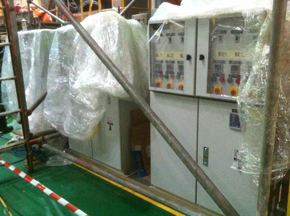 Electrical control panels wrapped in plastic, protected by scaffolding on a factory floor.
