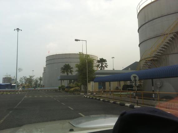 Large industrial tanks and structures in a gray, overcast setting; road and parked vehicles in foreground.