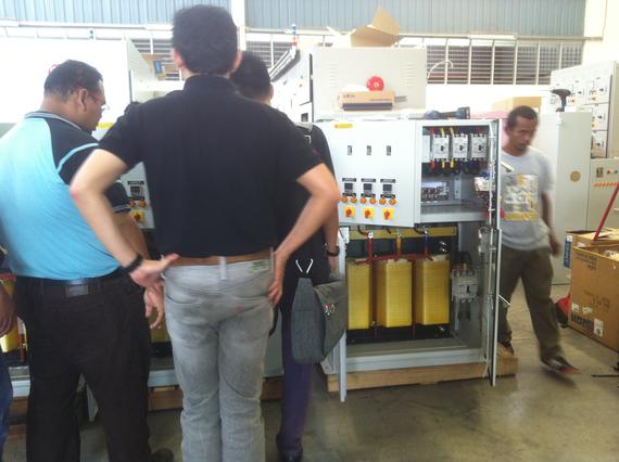 Group of people inspecting electrical equipment with yellow components in a factory setting.