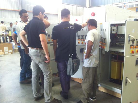 Group of men inspecting electrical panel in a warehouse; some are pointing and discussing.