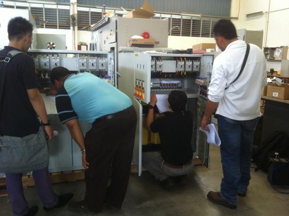 Four men examine an electrical panel with open doors, inside a workshop.