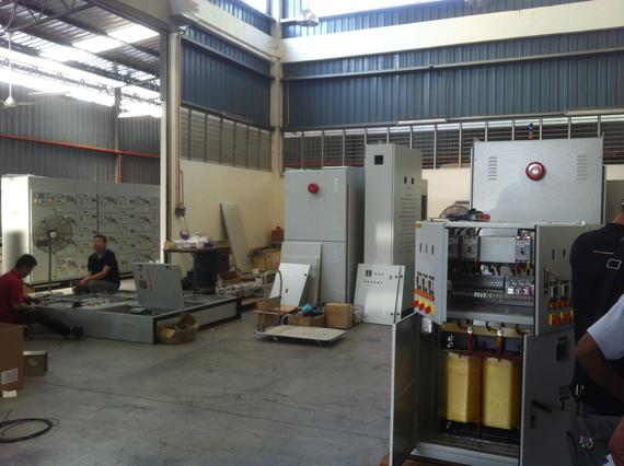 Inside a workshop, men assembling electrical panels. Gray metal boxes, open components, and equipment.