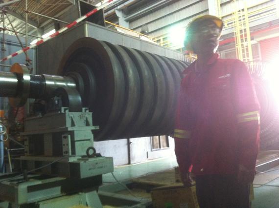 Man in red uniform beside a large industrial roller. Indoors, factory setting.
