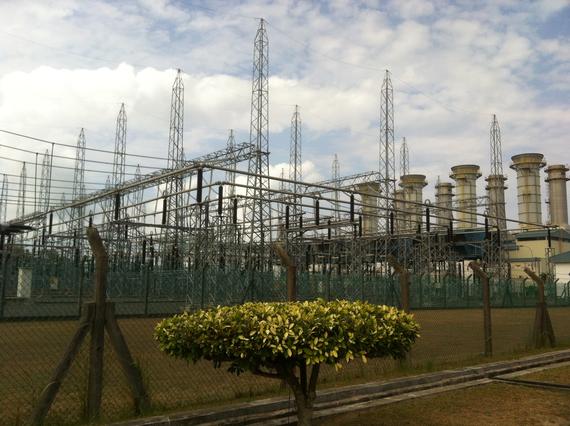 Electrical power plant with tall metal towers, chimneys, and a small tree in front.
