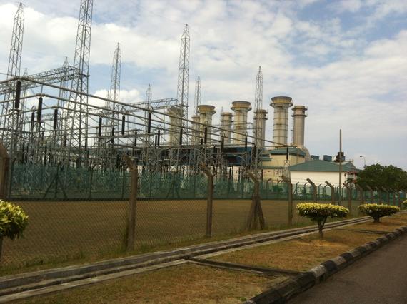 Power plant with electrical towers and chimneys under a cloudy sky. Green fence and brown grass.