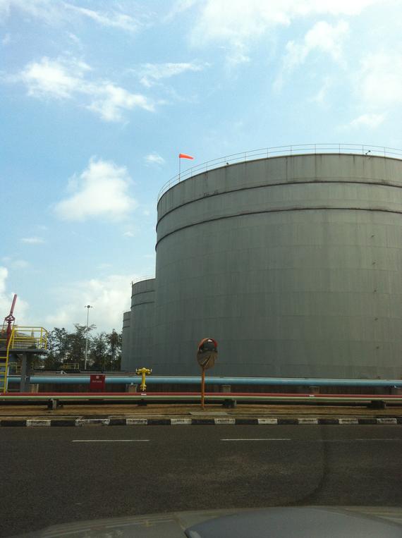 Large gray industrial storage tanks under a blue sky, with a road and some pipes in the foreground.