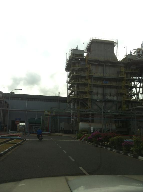 Industrial plant exterior with tall structures, scaffolding, and piping, with a person on a motorcycle in the foreground.