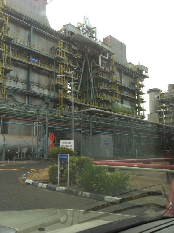 Exterior of a power plant with multiple levels, pipes, and walkways; a sign and vegetation in the foreground.