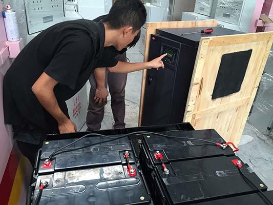 Two people examining a black safe with a control panel, connected to black batteries in an indoor setting.