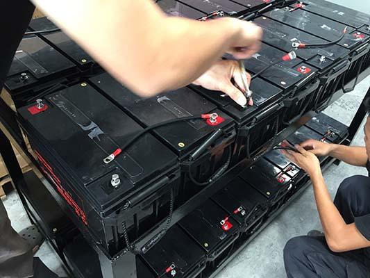 Two people connecting wires to a stack of black batteries with red terminals in a warehouse.