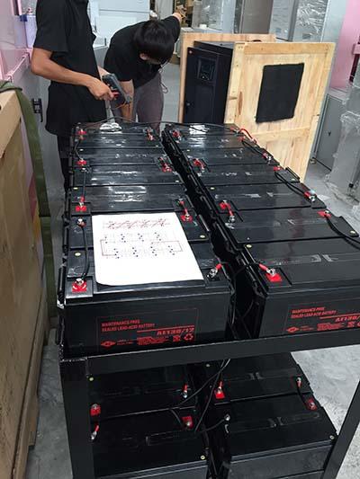 Two people working with a large array of black batteries on a metal shelf in an industrial setting.