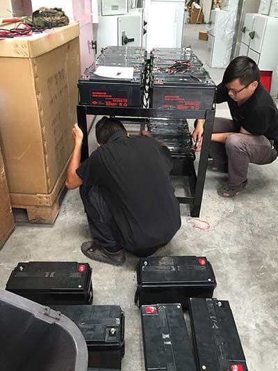 Two people installing batteries on a metal rack in a warehouse setting.