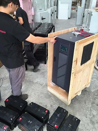 Two people moving a large dark appliance in a wooden crate, surrounded by batteries, in a factory setting.