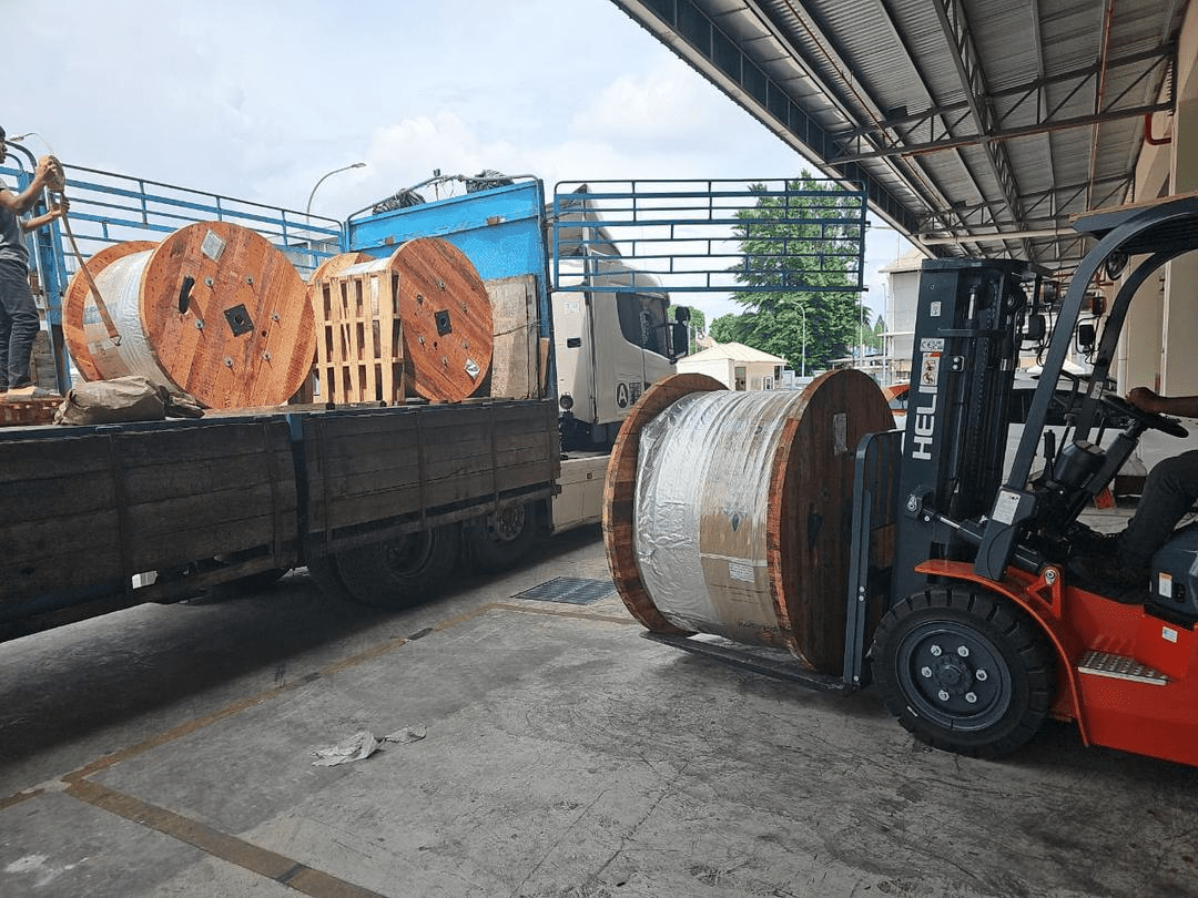 A forklift unloading large cable spools from a truck at a warehouse, under a cloudy sky.
