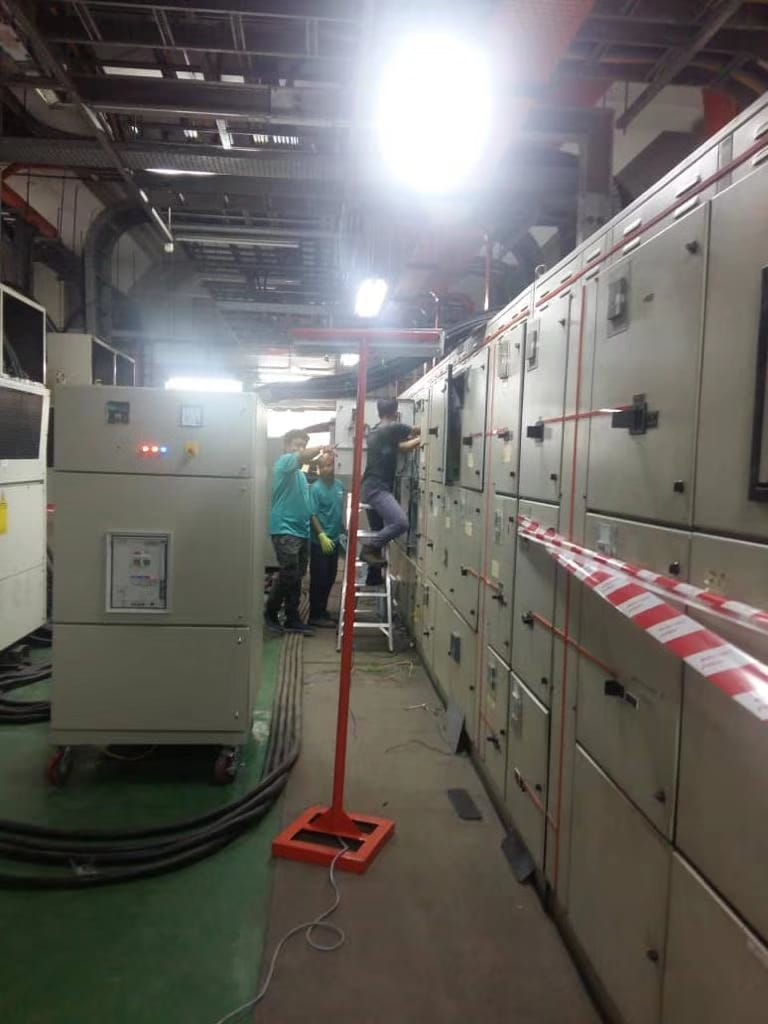 Electricians working on electrical panels in an industrial setting, illuminated by a work light.
